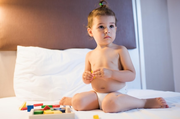 Portrait of adorable toddler beautiful girl siting on the bed in a toy mess playing with colorful construction wooden blocks in the lamplight Education game for kid in bedroom Nursery creativity