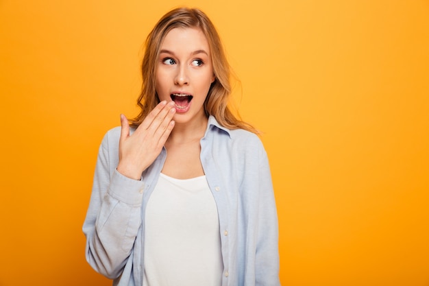 Portrait of adorable woman wearing braces looking aside and covering mouth with hand in surprise, isolated over yellow background