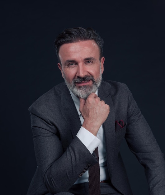 Portrait of adult businessman wearing trendy suit and sitting in modern studio on stylish chair against the black background. Horizontal mockup. High quality photo