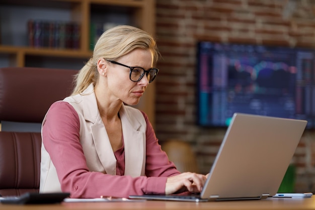 Portrait of adult businesswoman working with laptop at office Female trader texting messages doing paperwork with stock market graphs on background The rise and fall of cryptocurrencies concept