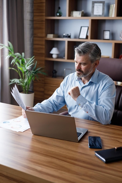 Portrait of adult grayhaired bearded businessman working with laptop at home office Texting messages doing paperwork calculating monthly expenses Business concept