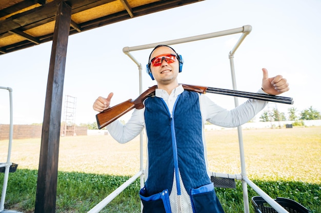 Portrait of adult smilling man in sunglasses protective headphones and a rifle vest with shotgun