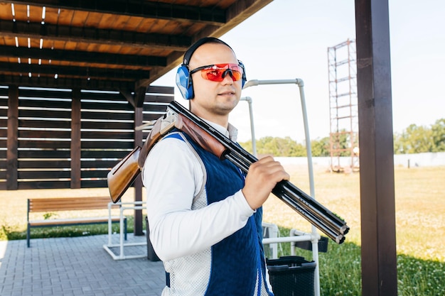 Portrait of adult smilling man in sunglasses protective headphones and a rifle vest with shotgun