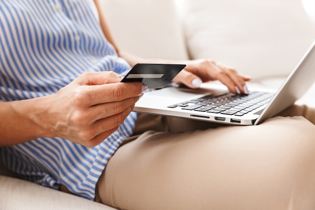 Portrait of adult woman in elegant clothes using laptop and credit card while sitting on couch in bright apartment