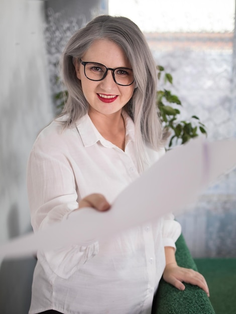 Portrait of an adult woman entrepreneur with grayish hair in a managerial position dressed in a white blouse and busy working in the office