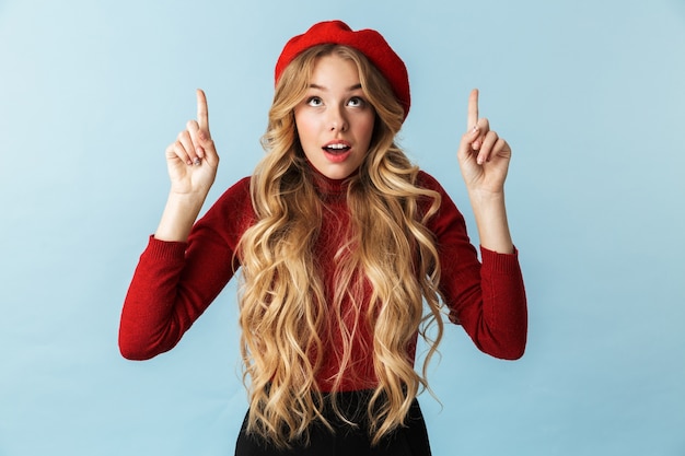 Portrait of affable blond woman 20s wearing red beret pointing fingers upward while standing, isolated 