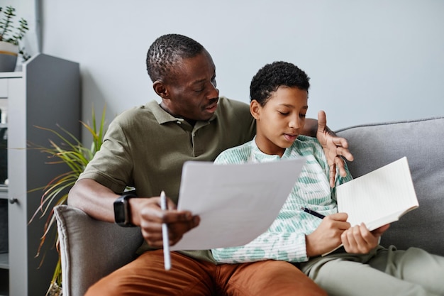 Portrait of african american father helping daughter doing homework while sitting on sofa at home co...