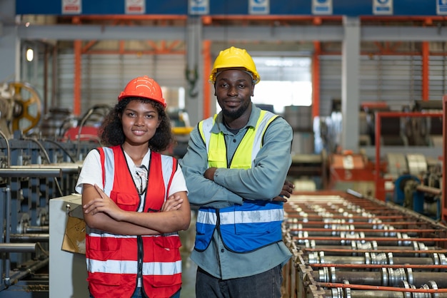 Portrait of African American female and male engineer worker wearing safety vest with helmet smiling arms crossed and standing look at camera while working in factory