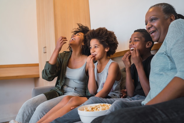 Portrait of African American grandmother, mother and children watching a movie and eating popcorn while sitting on sofa at home. Family and lifestyle concept.