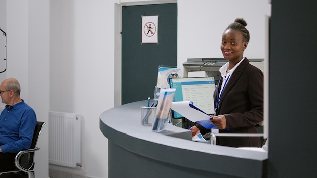 Portrait of african american receptionist working at registration desk in hospital waiting lobby. Female medical worker using checkup report forms on computer at reception counter.