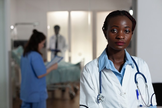 Portrait of african american woman with doctor occupation in hospital ward. Black surgeon, medic looking at camera with stethoscope, multi ethnic working team and patient at clinic