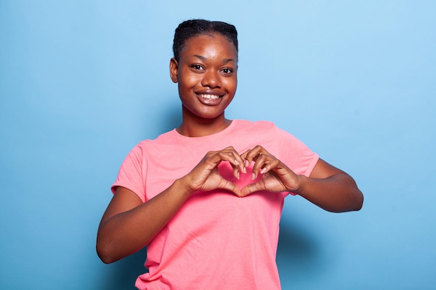 Portrait of african american young woman smiling at camera making heart shape symbol standing in studio with blue background. Friendly teenager showing affection. Valentines day concept