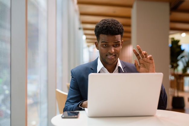 Portrait of African businessman using laptop computer in coffee shop while waving and having video call, horizontal shot