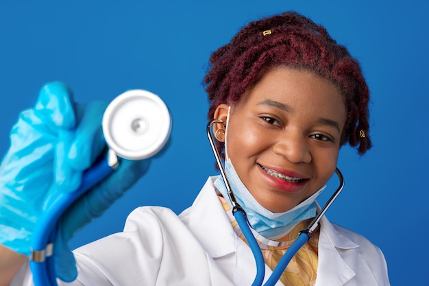 Portrait of african female doctor in lab coat with face mask and stethoscope against blue background