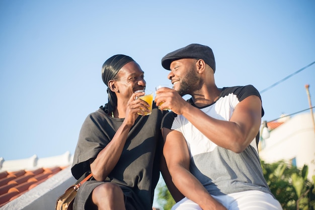 Portrait of African gay couple with glasses of juice. Two happy young men sitting together on roof top holding clinking glasses of juice looking at each other. Same sex couples love, relations concept