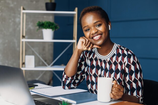 Portrait of an african woman posing at the office desk and looking at the camera while holding a cup...