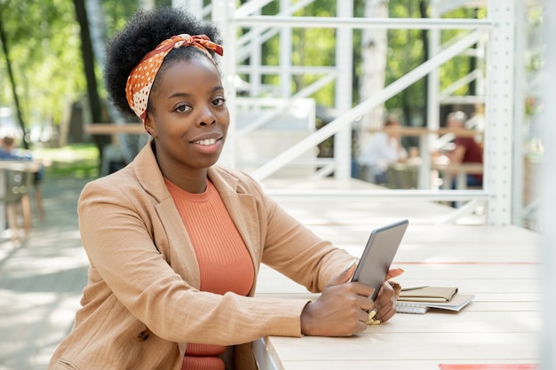 Portrait of African woman smiling at camera while sitting at the table in cafe and using digital tablet