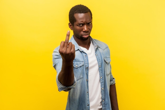 Portrait of aggressive hater angry man in denim casual shirt showing fuck sign with middle finger rude gesture of disrespect looking at camera indoor studio shot isolated on yellow background