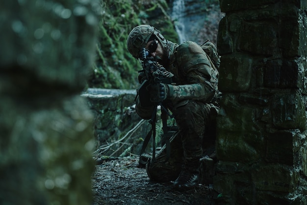 Portrait of airsoft player in professional equipment in helmet aiming at victim with gun in the forest. Soldier with weapons at war