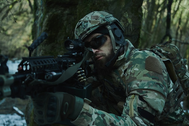 Portrait of airsoft player in professional equipment in helmet aiming at victim with gun in the forest. Soldier with weapons at war