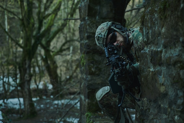 Portrait of airsoft player in professional equipment in helmet aiming at victim with gun in the forest. Soldier with weapons at war