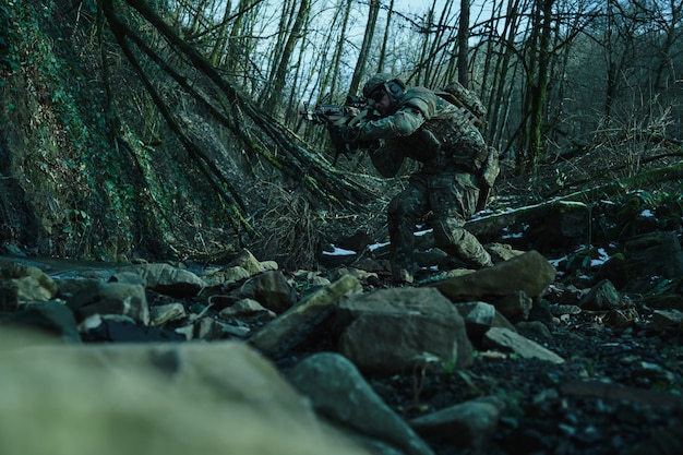 Portrait of airsoft player in professional equipment in helmet aiming at victim with gun in the forest. Soldier with weapons at war