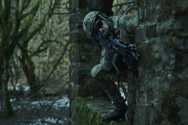 Portrait of airsoft player in professional equipment in helmet aiming at victim with gun in the forest. Soldier with weapons at war