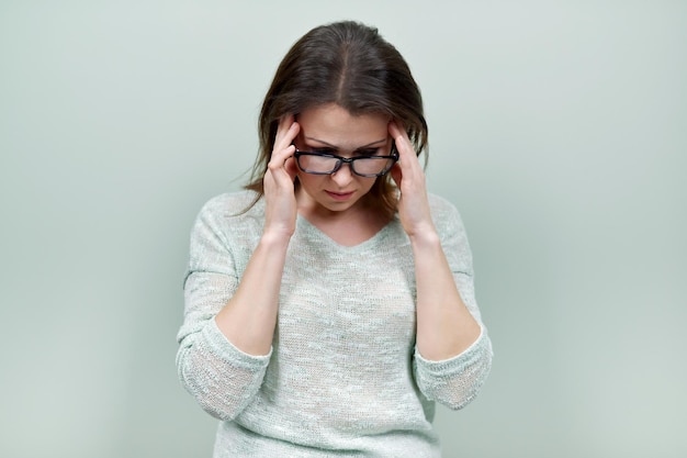 Portrait of alone sad mature woman with glasses, holding her head looking down, on green background. Negative human emotions, facial expressions, feelings, life perception