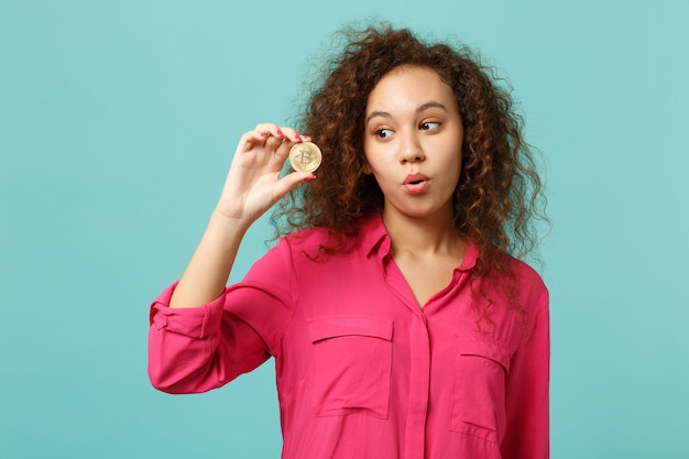 Portrait of amazed african girl in casual clothes holding bitcoin, future currency isolated on blue turquoise wall background in studio. People sincere emotions lifestyle concept. Mock up copy space.