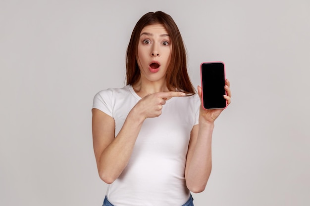 Portrait of amazed attractive young woman pointing at cell phone and looking with surprised expression shocked by mobile tariffs wearing white Tshirt Indoor studio shot isolated on gray background