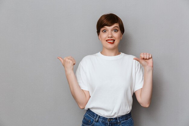 Portrait of amazed brunette woman wearing casual t-shirt pointing fingers aside at copyspace isolated over gray wall