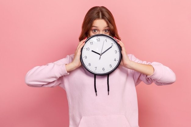 Portrait of amazed curly haired teenage girl in hoodie peeping behind big wall clock, time management and deadline. Indoor studio shot isolated on pink background