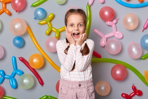 Portrait of amazed extremely happy birthday little girl with braids wearing casual clothing standing against gray wall with colorful balloons being pleasantly surprised