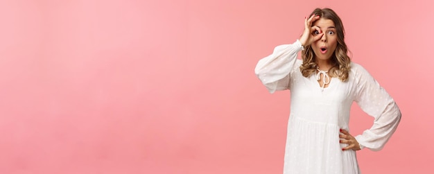 Portrait of amazed and intrigued young blond girl seeing something awesome look from okay sign with startled excited expression fold lips say wow pop eyes at camera pink background