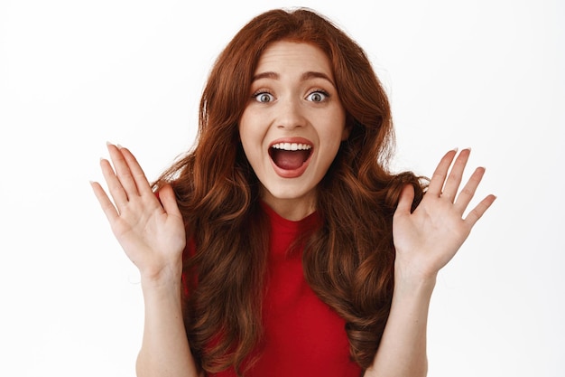 Portrait of amazed surprised redhead girl, staring astonished at camera, being fascinated by something awesome and super cool, standing against white background.