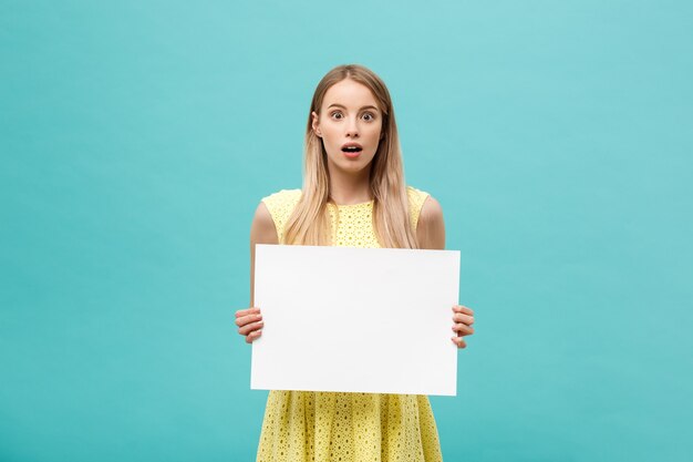 Portrait of amazed young blond woman holding blank sign with copy space on blue studio background. Showing shocked surprise face.