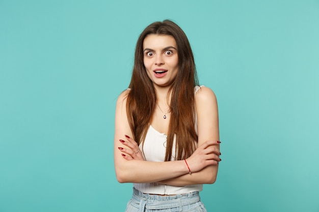 Portrait of amazed young woman in light casual clothes looking camera, holding hands crossed isolated on blue turquoise wall background. People sincere emotions, lifestyle concept. Mock up copy space.