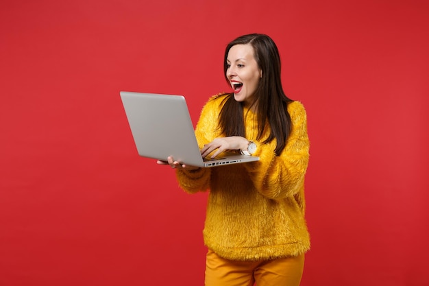 Portrait of amazed young woman in yellow fur sweater keeping mouth wide open using laptop pc computer isolated on bright red background. People sincere emotions, lifestyle concept. Mock up copy space.