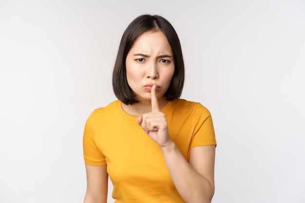 Portrait of angry asian woman shushing press finger to lips taboo stop talking sig looking annoyed scolding standing over white background