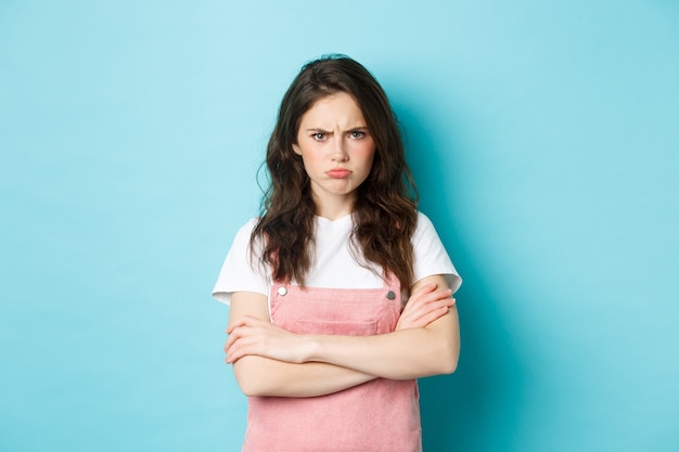 Photo portrait of angry and upset sulking girl, pouting about unfair situation, frowning and staring at camera, cross arms on chest defensive, standing over blue background.