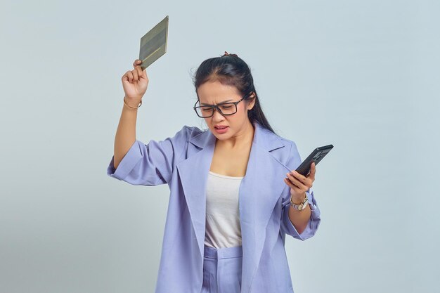 Portrait of angry young Asian woman with gesture of throwing away vehicle book and holding smartphone isolated on white background