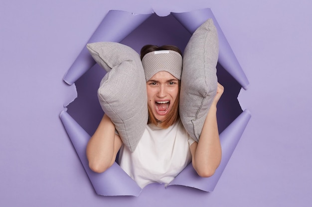 Portrait of annoyed sad angry young girl breaking through paper hole of purple background hearing loud noise in early morning being in bad mood covering ears with pillows