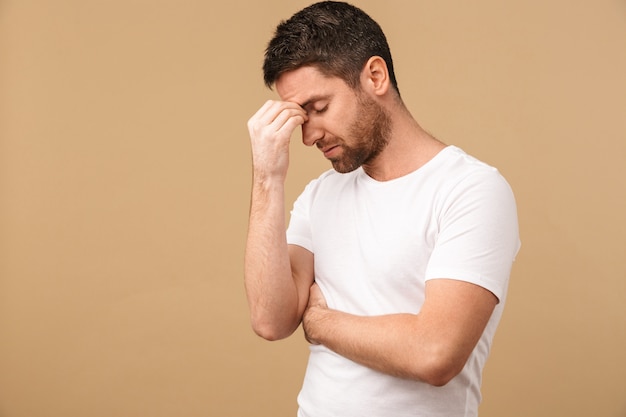 Portrait of an annoyed young man casually dressed standing isolated over beige, having a headache