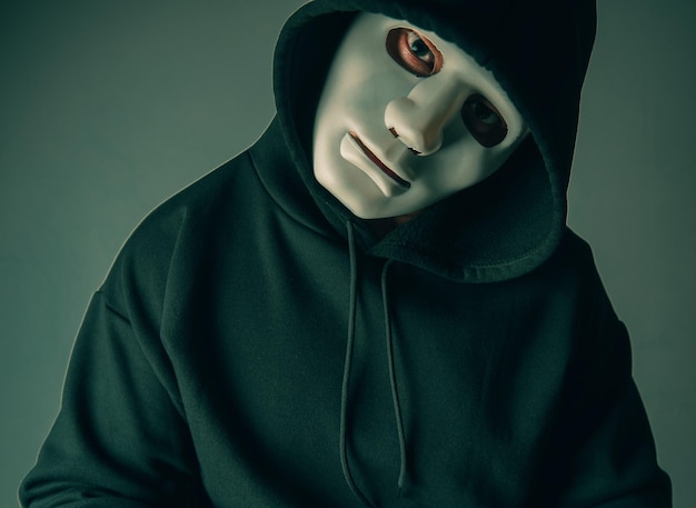 A portrait of an anonymous hacker wearing a mask and a black hoodie sitting with his head tilted and terrifying Hacking and malware concept