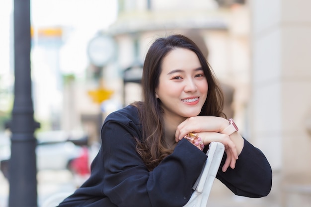 Portrait Asian beautiful female in dark blue long sleeve shirt sits and smile on chair in an urban outdoor park.