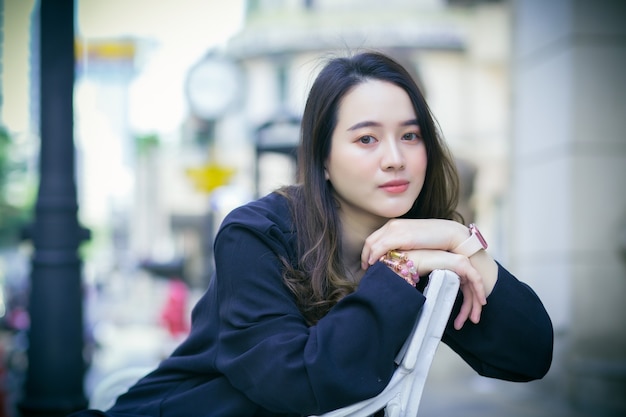 Portrait Asian beautiful woman in dark blue long sleeve shirt sits and smile on chair in an urban outdoor park.