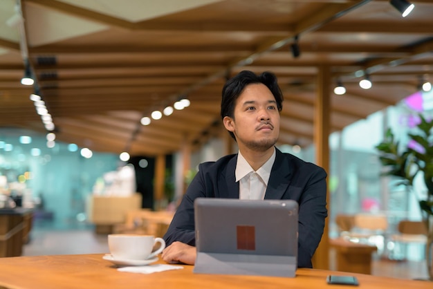 Portrait of Asian businessman in coffee shop thinking and using digital tablet computer