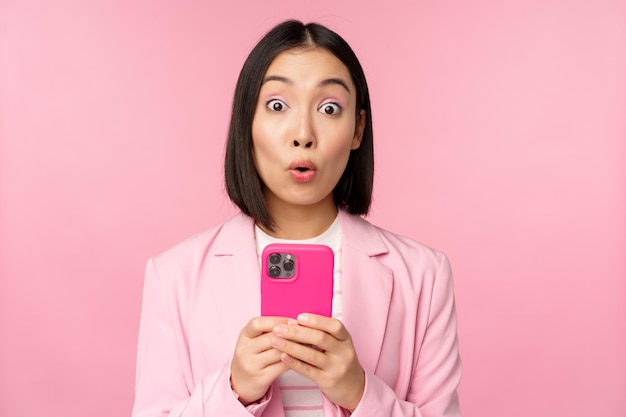 Portrait of asian businesswoman with surprised face using smartphone app wearing business suit Korean girl with mobile phone and excited face expression pink background