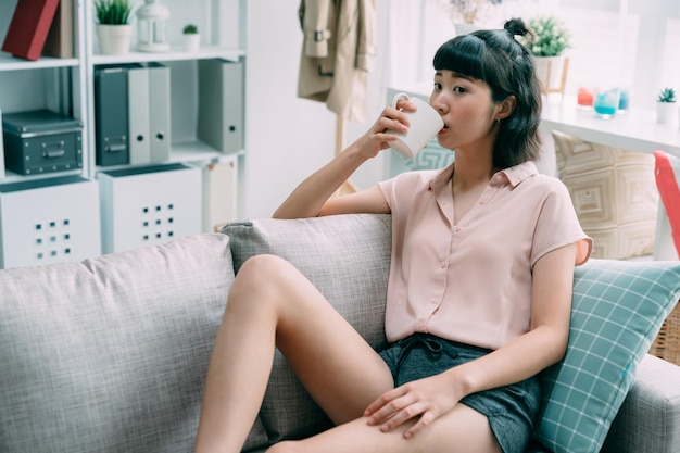 Portrait of asian chinese charming woman holding cup of tea looking away. young girl leaning lying on comfort sofa drinking beverage. cute female enjoy coffee in morning sitting on couch in cozy home