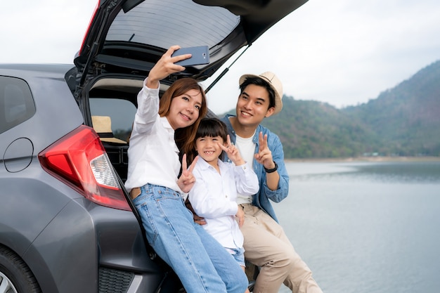 Portrait of Asian family sitting in car with father, mother and daughter selfie with lake and mountain view by smartphone while vacation together in holiday. Happy family time.